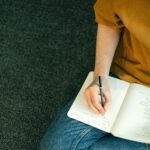 Close-up of a person taking notes in a notebook while sitting on the floor, emphasizing education and focus.