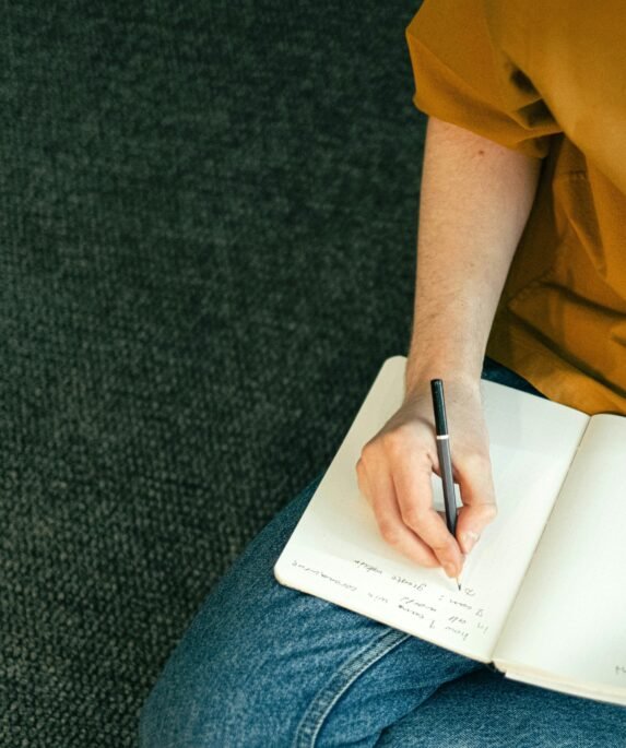 Close-up of a person taking notes in a notebook while sitting on the floor, emphasizing education and focus.
