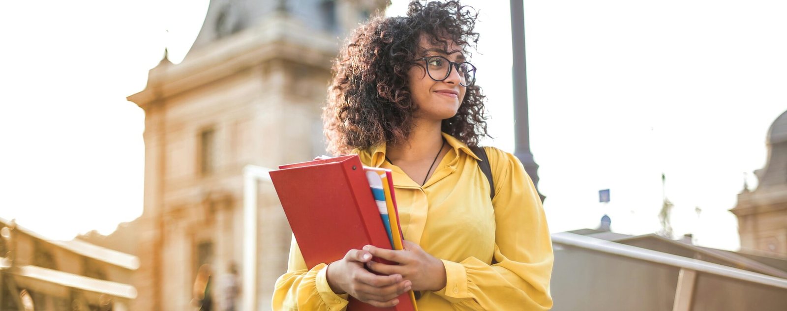 Smiling female student with curly hair and glasses holding books on campus steps.