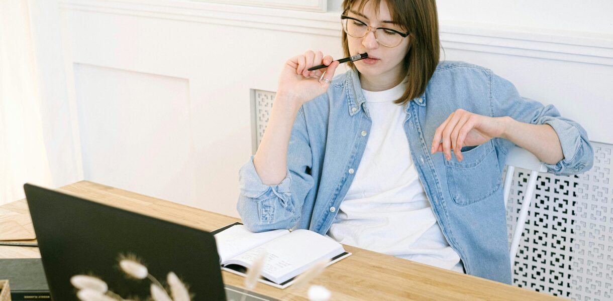 Woman working thoughtfully in home office, reflecting on ideas with laptop and notebook.