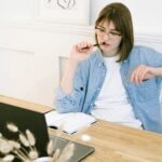 Woman working thoughtfully in home office, reflecting on ideas with laptop and notebook.