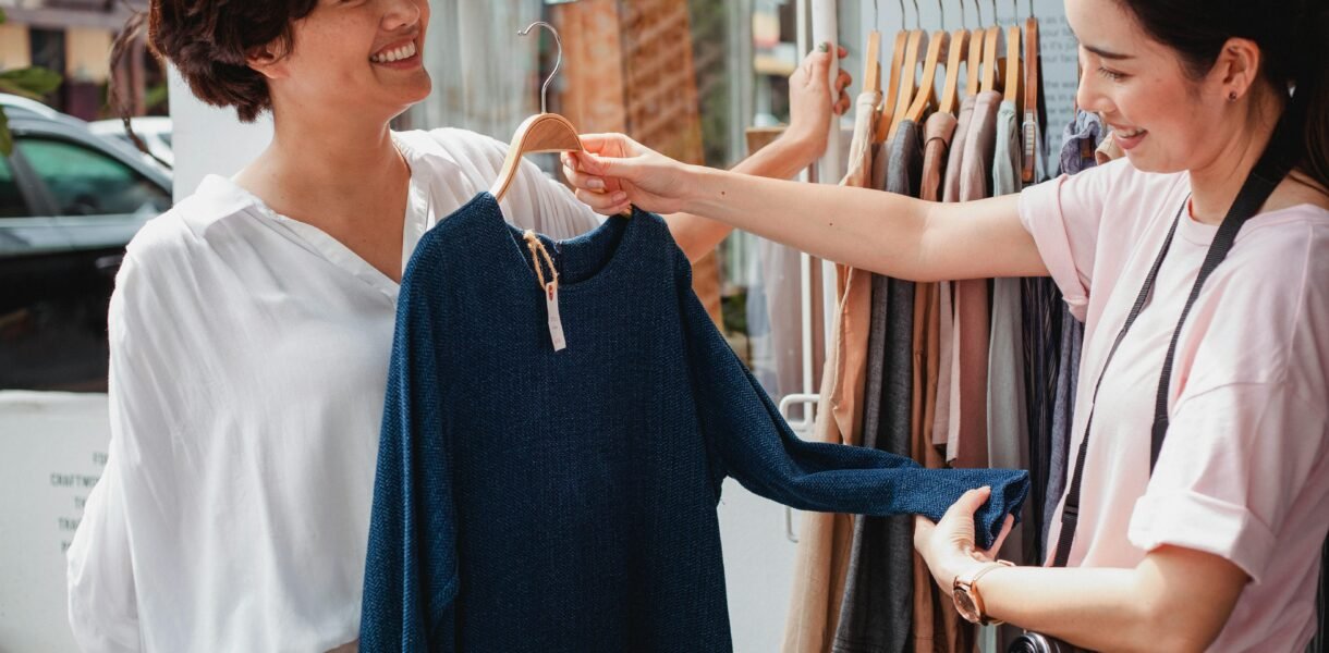 Two women smiling while shopping for clothes outdoors on a sunny day.