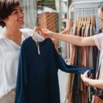 Two women smiling while shopping for clothes outdoors on a sunny day.