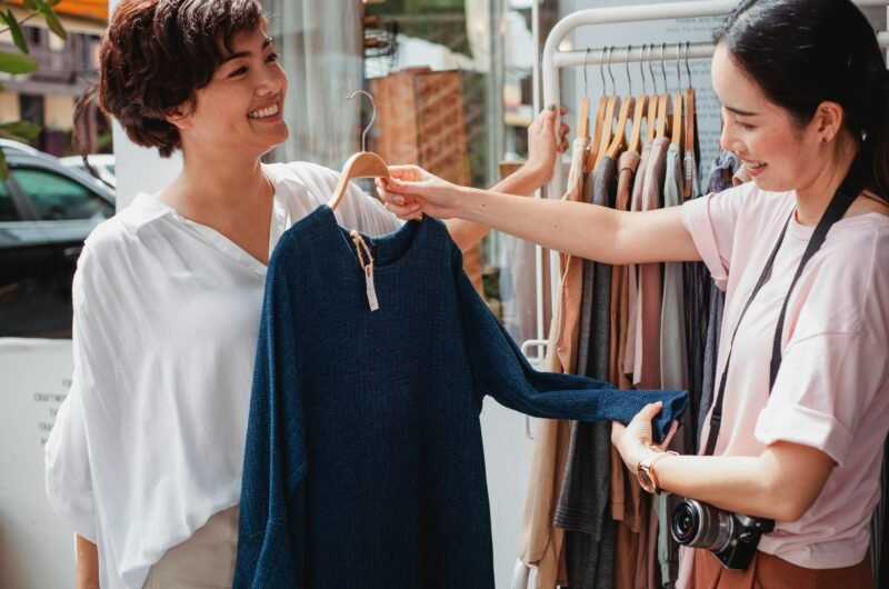 Two women smiling while shopping for clothes outdoors on a sunny day.