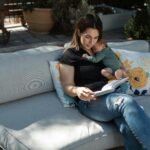 A serene moment of a mother reading while holding her sleeping baby on an outdoor sofa.