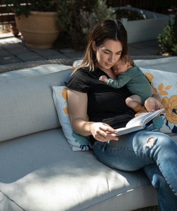 A serene moment of a mother reading while holding her sleeping baby on an outdoor sofa.