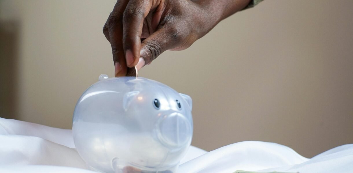Close-up of a person's hand placing coins into a transparent piggy bank to save money.